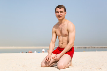 A man is kneeling in the sand. A resting European in red shorts knelt on the beach.
