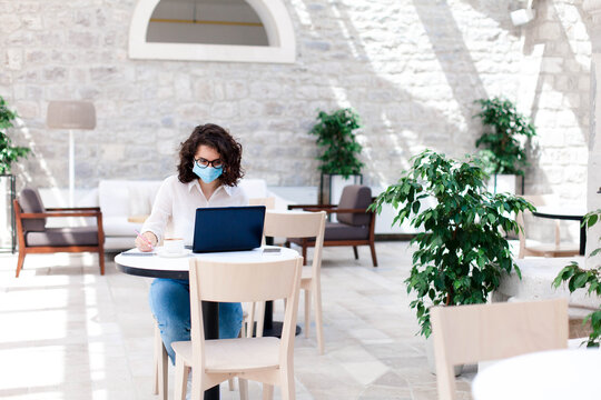 Young Woman Working Safety In Cafe Indoors. Social Distancing During Quarantine. Freelancer Wearing Protective Mask, Using Laptop. Comfortable Office, Coworking Modern Workplace With Limited Seating.