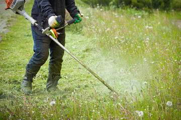 Worker in dark protective clothing with a gas mower in his hands, mowing grass in front of the house. A man mows grass with dandelions on a rainy spring day. Trimmer in the hands of a man.