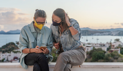 Two young women friends communicate have fun at sunset on the roof with a smartphone, with protective masks on their faces