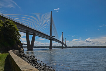 Queensferry Crossing over River Forth in Scotland, UK. Taken on a sunny, summer day with blue sky and light clouds. Sea wall, rocks, grass and trees in foreground.