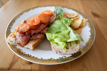 Homemade bacon and fried tomato and lettuce baguette open sandwich, Sometimes called a BLT before closing on a white plate with a gold trim with crisps. On a wooden table, lit with natural light