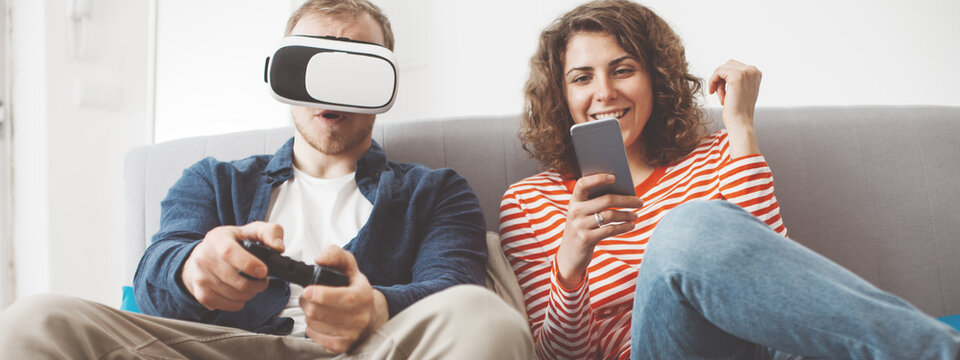 Happy family living together with electronic devices. Man playing breathtaking console game with VR headset. Woman typing message using mobile phone