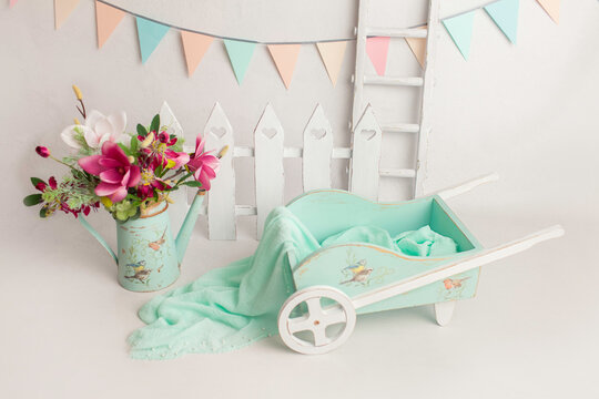 Green spring cart with birds near the fence and stairs. watering can with a floral bouquet in white baby's room, copy space concept. interior of children's room. Cute baby girl sitting in a trolley.