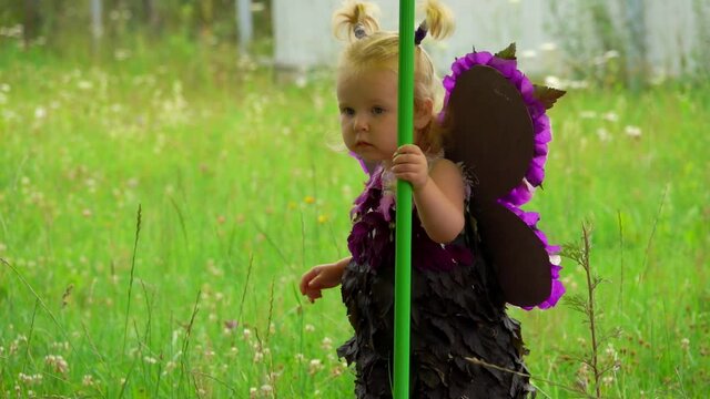 Little cute blond girl with purple butterfly wings walks around green pole outdoors