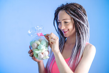 A young happy woman with dreadlocks is holding a package of multi colored meringues on a blue background. Concept of a bright summer mood