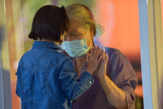 A Sad Hand Of Grandchild And Grandmother Mature Woman In A Respiratory Mask Communicates Through A Window. Elderly Quarantined, Isolated. Visit To Grandma.selective Focus