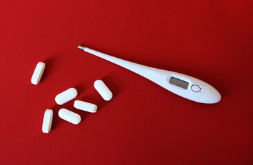 Oval white biconvex tablets and white digital thermometer on red background with light shadows....