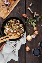 Roast forest mushrooms, potatoes, onions and seasonal herbs in a cast iron pan and ingredients on a wooden background outdoors.
