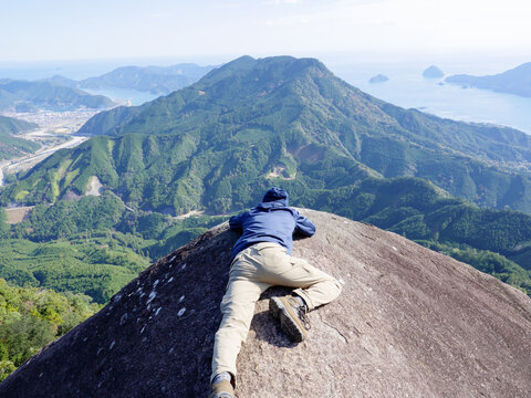 三重県尾鷲市にある便石山の像の背。恐る恐る絶壁の先から寝そべりながら景色を見る男性

