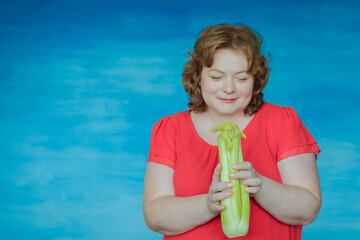 Cute smiling chubby red-haired girl bites celery and smiles against a blue background.