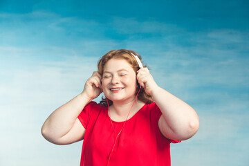 Young attractive plump woman with red hair listening to music with pleasure headphones on a blue...