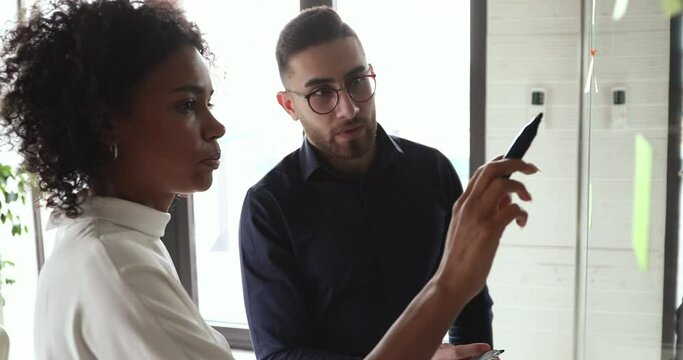 Two diverse team people brainstorm draw on glass wall discuss creative strategy at office meeting. African mentor coach teaching arab employee collaborating in research startup teamwork concept.