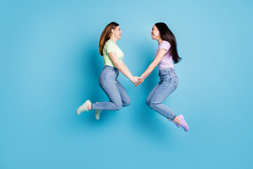 Fototapeta na wymiar Full body profile photo of two people lesbians couple ladies jump high up hold hands good mood rejoicing happy together wear casual t-shirts jeans footwear isolated blue color background