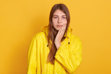 Upset female wearing yellow jacket touching her cheek, suffering from toothache, looks at camera, having medical problems, standing against yellow wall.