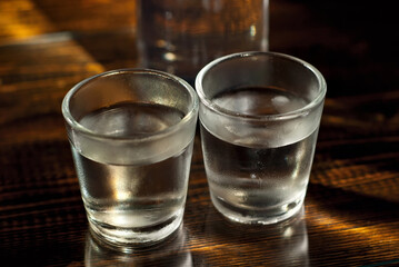 Frozen glasses with alcohol near the bottle. Drinks on a dark brown table. Transparent utensils on a black background. Old shabby boards under glass.