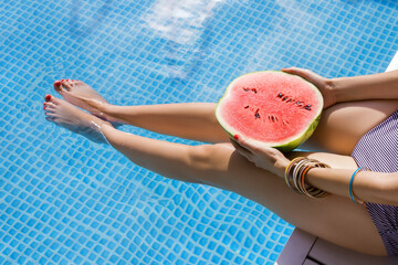 Top view female slim legs. Woman is relaxing, sunbathing, holding watermelon sitting on edge of pool. Girl in swimsuit is resting, lounging on resort hotel. Vacation, traveling, summer mood.