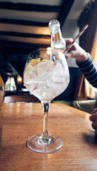 A beautiful refreshing sparkling gin and tonic alcohol alcoholic  beverage being poured with fresh lemon slices and big freezing cold ice cubes, in a traditional english pub on a wooden table.