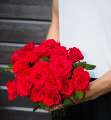 Male person holding a beautiful bouquet of red roses wearing white shirt and black pants against a gray wooden wall