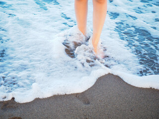 Unrecognizable female person walking alone in spuming sea water on a beach barefoot