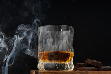 A glass of whisky together with dark chocolate on wooden surface against dark background and smoke