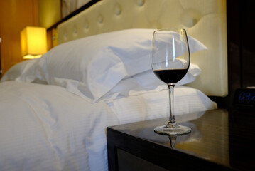 A glass with red wine stands on the bedside table. Bed with pillows and a lamp in the background.
