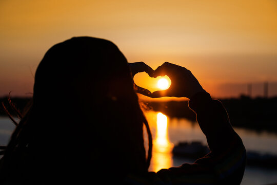silhouette of a girl and hands forming a heart shape with sunset and reflection of a solar track in the water. expression of love and gratitude to the world