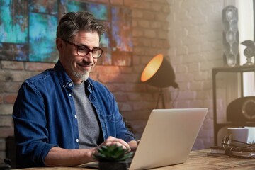 Portrait of handsome mature man with grey hair, wearing red glasses, working on laptop, sitting at desktop at home or in the office.