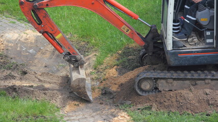 A compact crawler excavator clears paving stones in the Park with a bucket after works on a summer day on green grass background, landscape design