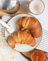 Continental breakfast, a cup of coffee with milk, pair of croissants on ceramic plate with butter and orange jam on a wooden cutten board, view from above, copy space
