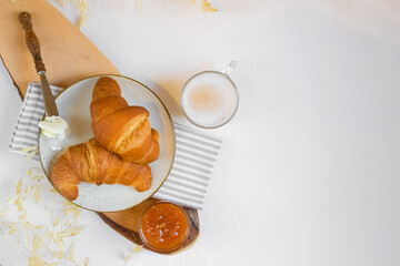 Continental breakfast, a cup of coffee with milk, pair of croissants on ceramic plate with butter and orange jam on a wooden cutten board, view from above, copy space