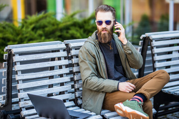 Young bearded hipster man sitting on the bench and talk phone outdoors