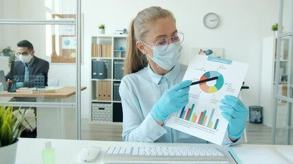 Portrait of young female employee making online video call from office talking and showing charts during infection epidemic, woman is wearing face mask and rubber gloves. - Powered by Adobe