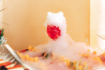 Fototapeta na wymiar Smoking glass at fruit and vegetable buffet table. Closeup of glass of red beverage with white dense fog. Chemical reaction of dry ice with water.