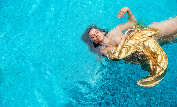 beautiful young proud woman in gold dress, evening dress floating weightlessly elegant floating in the water in the pool dark hair drifting in turquoise water, copy space, space for text,