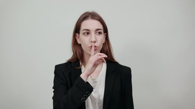 Index finger on lips, silence gesture, shhh quiet, asks for voicelessness forefinger at the mouth. Young attractive woman with brown hair in a light t-shirt and black jacket on white background