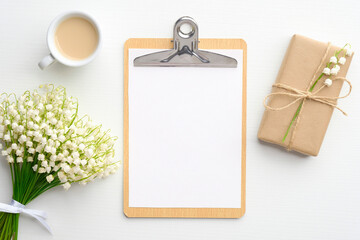 Wedding checklist concept. Clipboard with blank paper mockup, gift box wrapped craft paper, cup of coffee and lily of the valley flowers on white table. Flat lay, top view.