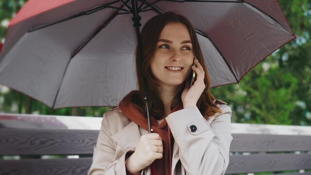 season, weather and people concept - young brunette woman sitting on a bench with an umbrella on a rainy day. woman talking on a modern phone, writing a message, woman looking at a mobile phone