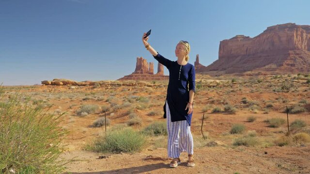 young woman in light white pants and a dark blue tunic dress on a background of red fancy mountains Monument Valley takes a selfie on a mobile phone camera life style freedom photo shoot