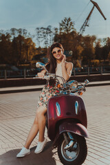 Fototapeta na wymiar Portrait of a young brunette in a colorful dress and sunglasses in the summer in the city on a scooter 