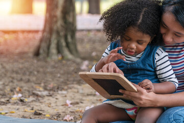 Close up young mother holding blackboards with her daughter while sitting on lawn learning something together in the park. Mixed race little curly hair girl sitting on knees of her mom.