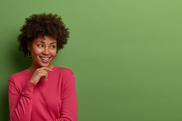 Fototapeta na wymiar Dreamy cheerful curly African American woman holds hand on chin, sees something pleasant and appealing, dressed in pink jumper, stands against green background, free space for your advertisement