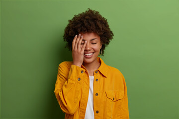 Fototapeta na wymiar Pretty joyful dark skinned woman keeps hand on face, laughs at funny joke, closes eyes from joy, expresses positive emotions, wears yellow fashionable jacket, poses indoor over green studio wall