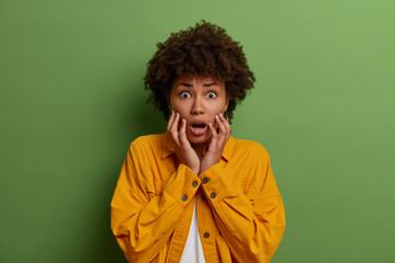 Portrait of astonished scared woman grabs face, stands alarmed as afraids of something, hears terrifyig bad news, stares with bugged eyes at camera, dressed in yellow jacket, isolated on green