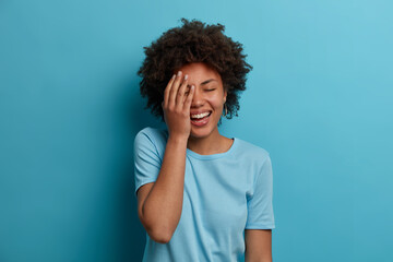 Obraz na płótnie Canvas Horizontal shot of happy dark skinned woman makes face palm, giggles positively, keeps eyes closed, has good mood, wears blue t shirt, poses indoor, hears amazing info, reacts to lucky fine situation