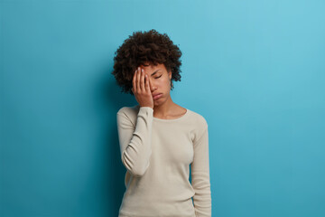 Fototapeta na wymiar Frustrated unhappy tired young woman makes face palm, keeps eyes closed and sighs from tiredness, wears white jumper, poses against blue background, bothered by something annoying, feels fed up