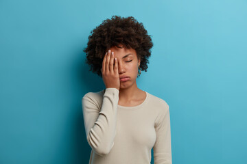 Fototapeta na wymiar Exhausted overworked female student makes face palm, closes eyes from tiredness, spend sleepless night preparing for exams, poses unhappy indoor, wears casual white jumper, isolated on blue wall