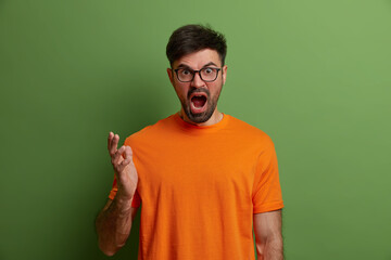 Outraged stressful European man screams from annoyance, gestures angrily, argues with someone, looses temper and feels aggression, shakes hands, wears orange t shirt, isolated on green background