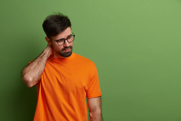 Sad fatigue man concentrated down, keeps hand on neck, has thoughtful expression, thinks how to solve problem, being in despair, dressed casually, poses over vivid green background, copy space