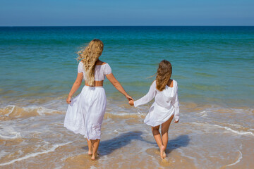 Young beautiful mother with her charming daughter in white clothes walk on a sandy beach near the water